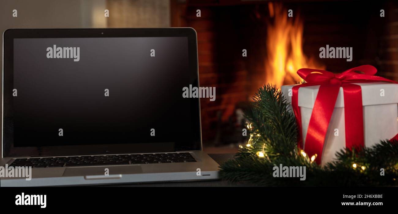 Regalo di Natale e schermo bianco laptop su sfondo camino acceso, concetto di casa vacanze invernali ufficio. Casa accogliente natale Foto Stock