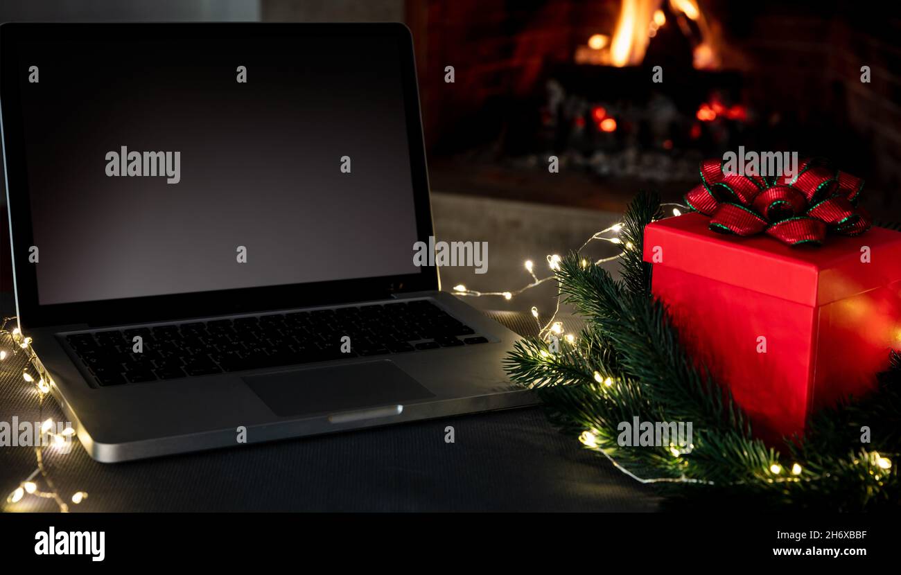 Computer portatile con schermo vuoto e regalo di Natale su sfondo camino acceso, concetto di casa vacanze invernali ufficio. Casa accogliente natale Foto Stock