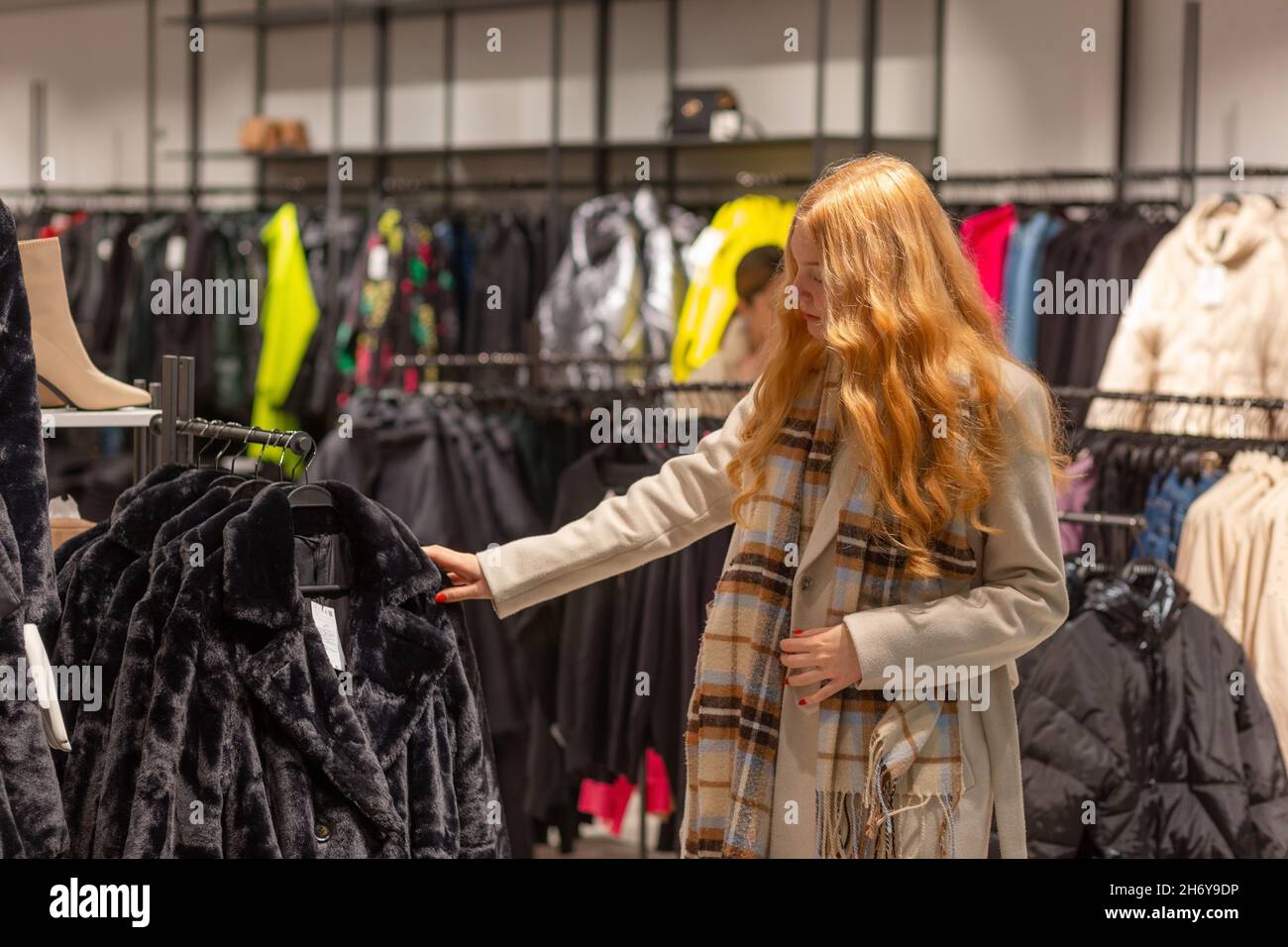 Giovane cliente femminile con i capelli dorati che esaminano i migliori cappotti di pelliccia in moderno negozio di stoffe di donne Foto Stock