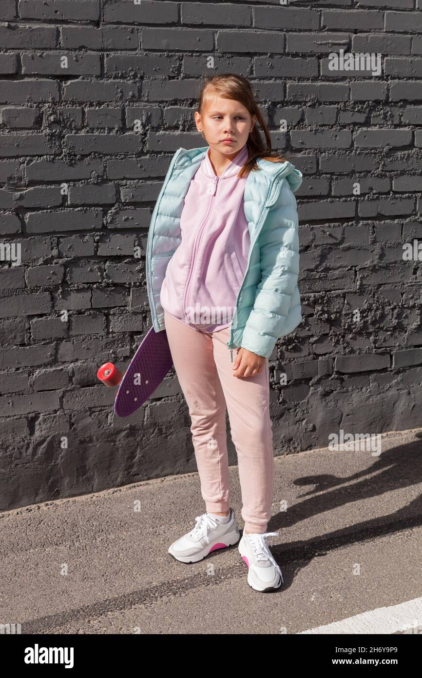 Bambina in abiti colorati si trova vicino a una parete di mattoni grigi con uno skateboard in mano Foto Stock