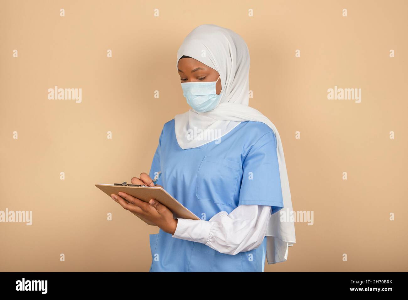 Una giovane infermiera musulmana in piedi in una maschera chirurgica prende nota su una clipboard su una parete gialla Foto Stock