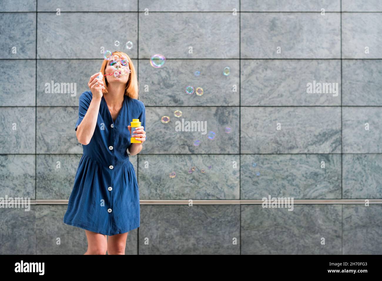 Spensierate la giovane donna in abito blu soffia le bolle di sapone arcobaleno mentre si diverte in estate su sfondo grigio parete Foto Stock