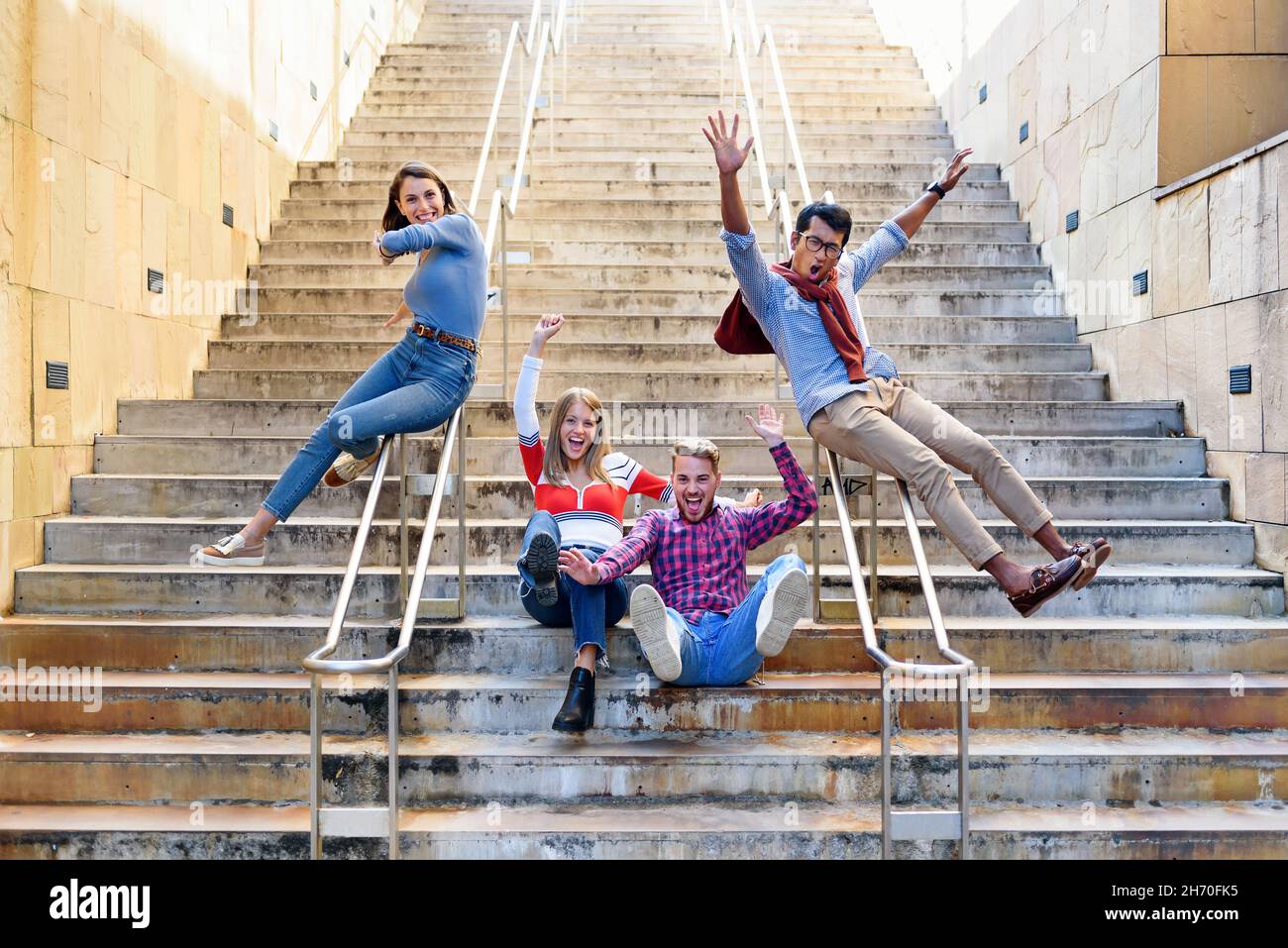 Un gruppo multiculturale di amici felici ed eccitati che si acclamano e ridono mentre scivolano sulle rotaie di una rampa di scale esterne in una città in libertà Foto Stock