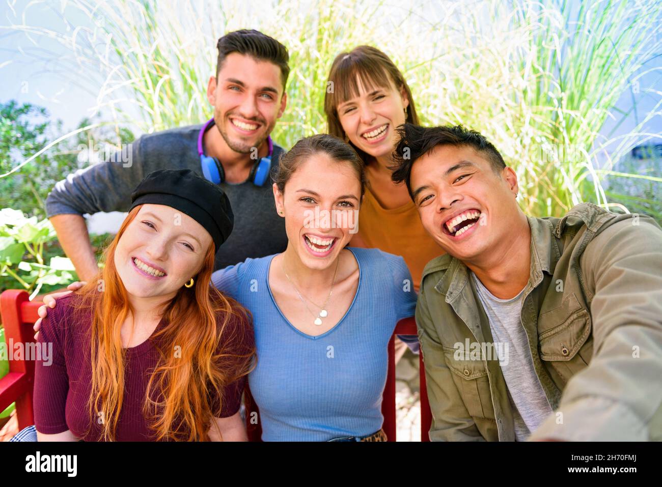 Gruppo di allegri amici multietnici maschi e femmine in abiti casual sorridenti e guardando la macchina fotografica mentre si prende selfie in giardino verde il giorno di sole Foto Stock