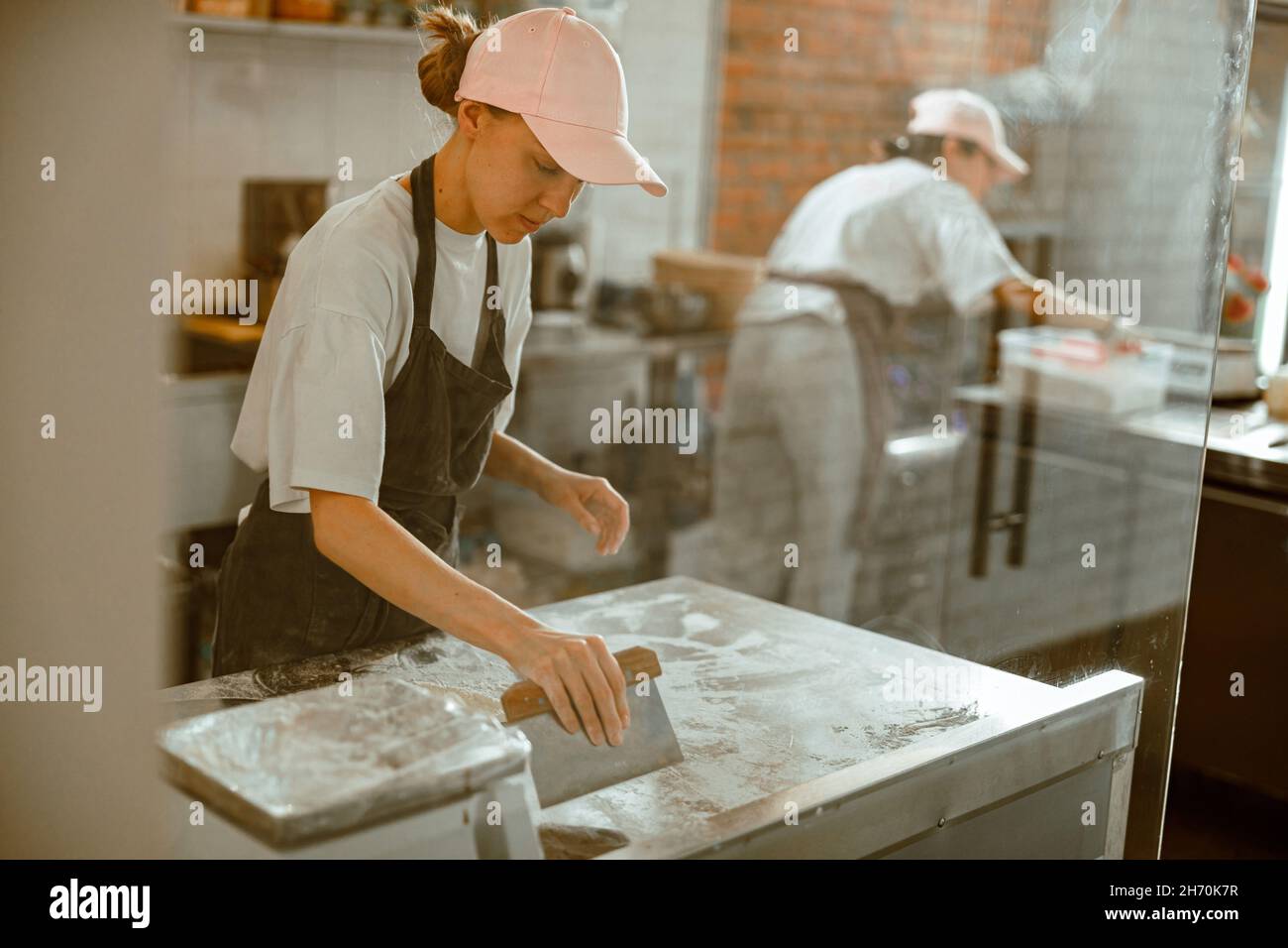 La donna graziosa pulisce il piano di lavoro dalla farina dopo aver preparato l'impasto in un laboratorio di panetteria Foto Stock