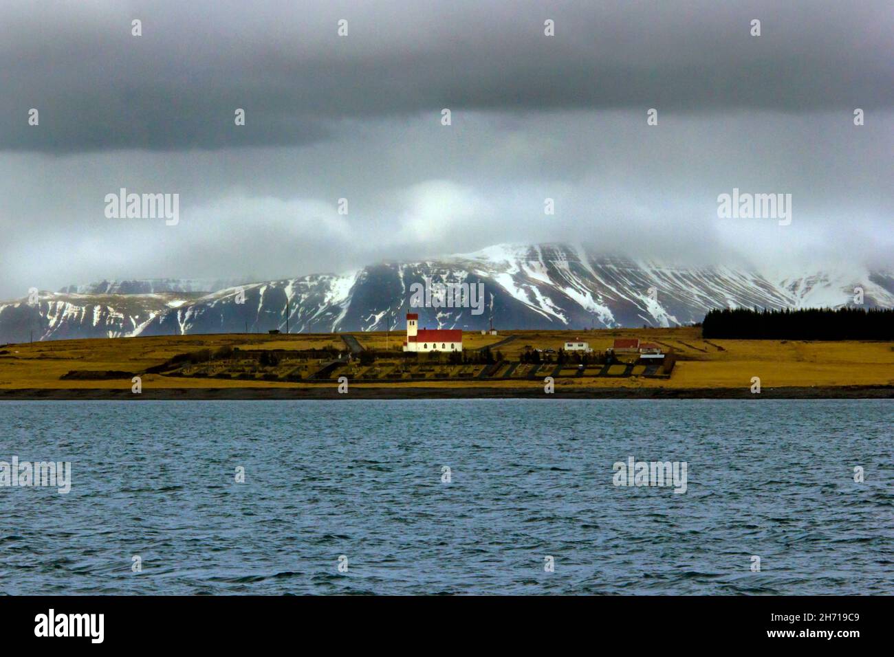 Chiesa rossa sul tetto in Islanda con il mare in primo piano montagne con neve e ghiaccio dietro spazio copia Foto Stock