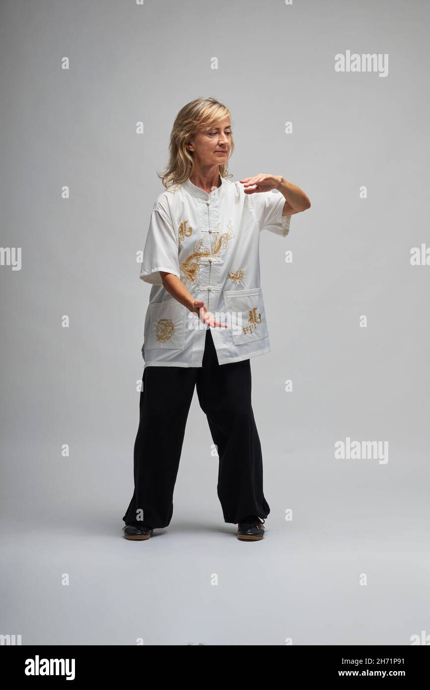 Donna bionda matura praticando Chi Kung e Tai Chi su sfondo bianco. Indossa una tradizionale giacca cinese Tai Chi bianca, pantaloni neri e bl Foto Stock
