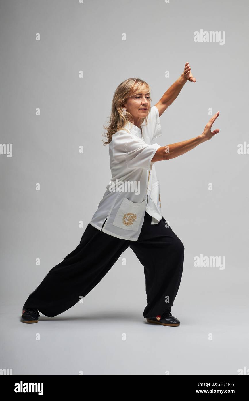 Donna bionda matura praticando Chi Kung e Tai Chi su sfondo bianco. Indossa una tradizionale giacca cinese Tai Chi bianca, pantaloni neri e bl Foto Stock