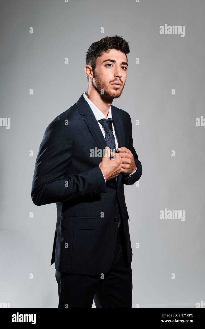 Giovane uomo con barba che indossa un abito blu scuro con la mano destra che tocca la giacca e guarda la fotocamera Foto Stock