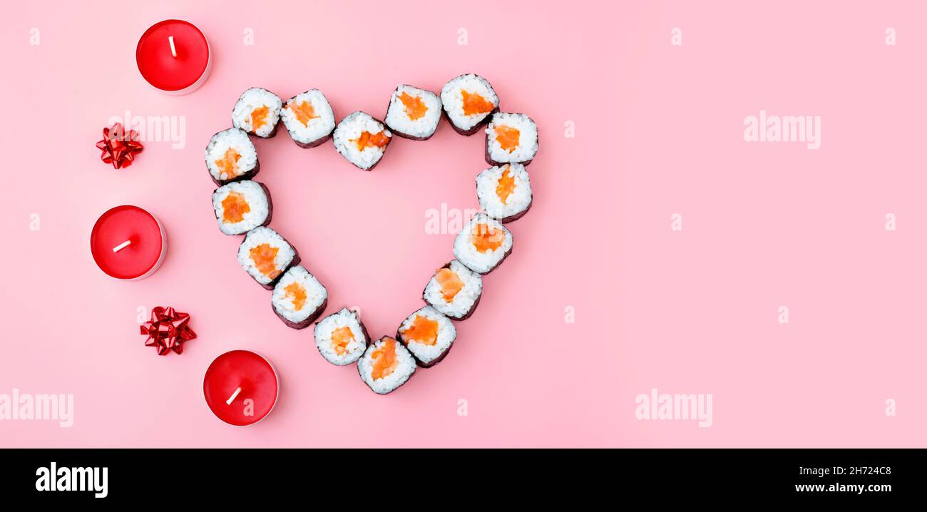 Sushi di salmone rotolini disposti a forma di cuore, accanto a candele rosse e bowson uno sfondo rosa. Il concetto di cucina giapponese per San Valentino Foto Stock