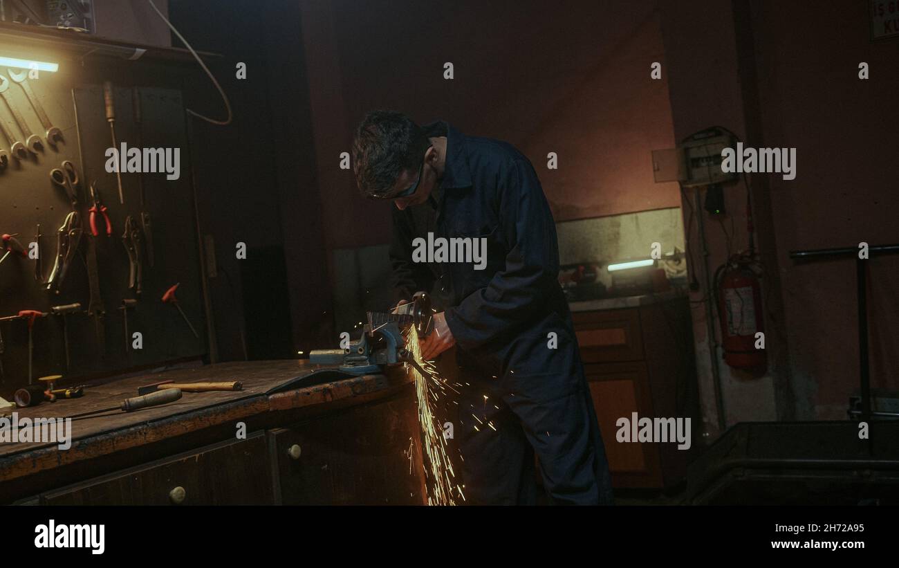 Il giovane uomo fabbro usa la macchina da taglio del ferro o del metallo e la macinazione del metallo sul pezzo di ricambio dell'acciaio in officina Foto Stock