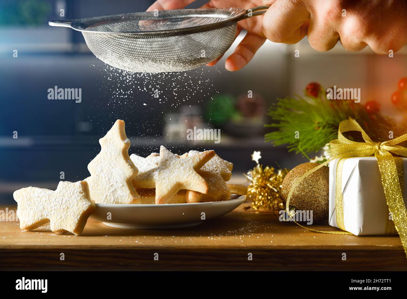 Cospargere i biscotti natalizi fatti in casa su un piatto sulla panca della cucina con zucchero a velo e un setaccio da cucina. Vista frontale. Composizione orizzontale. Foto Stock