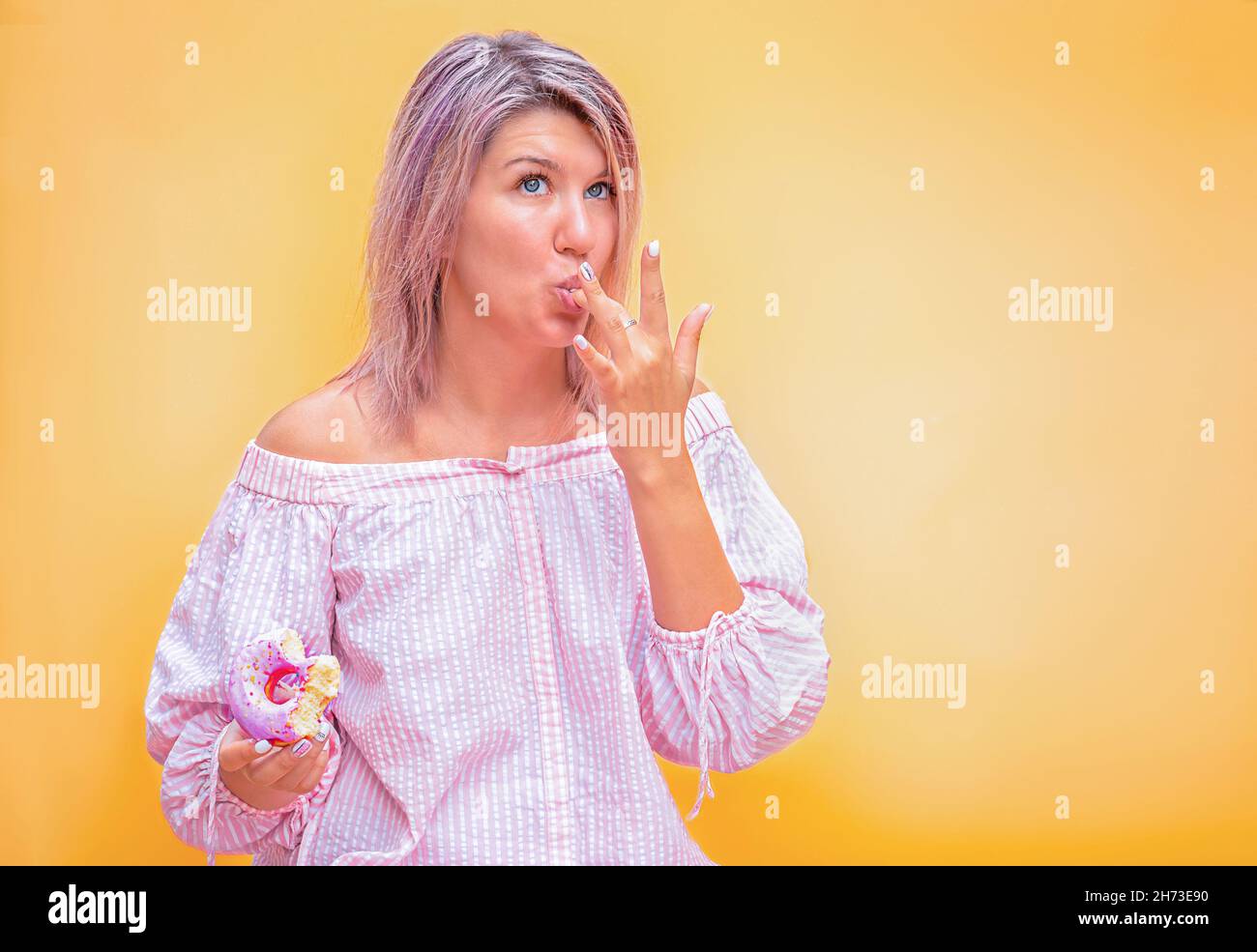 la giovane donna lecca la glassa dalle dita, d'altra parte una ciambella rosa su sfondo giallo Foto Stock