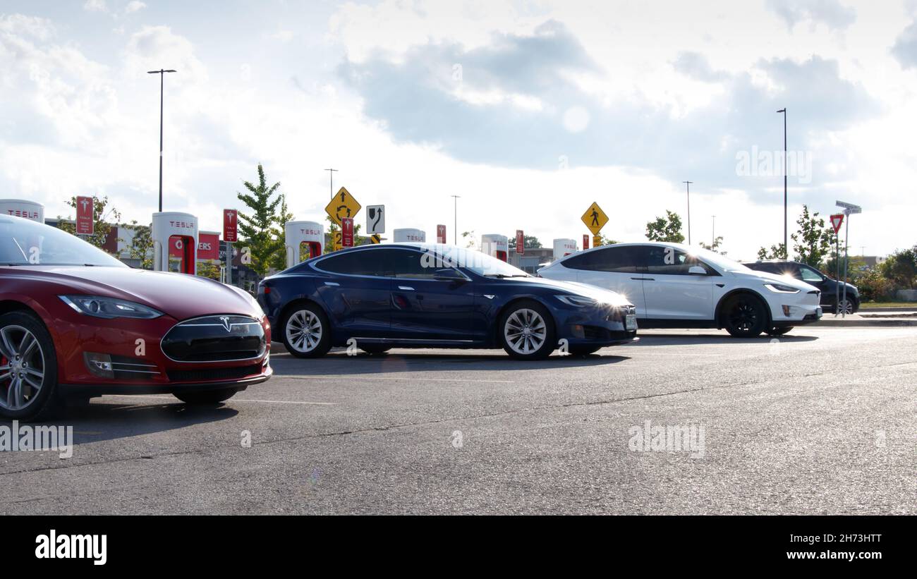 Una Tesla Model S rossa e blu e una Model X bianca vengono visualizzate durante il giorno parcheggiate presso una stazione Supercharger Tesla. Foto Stock