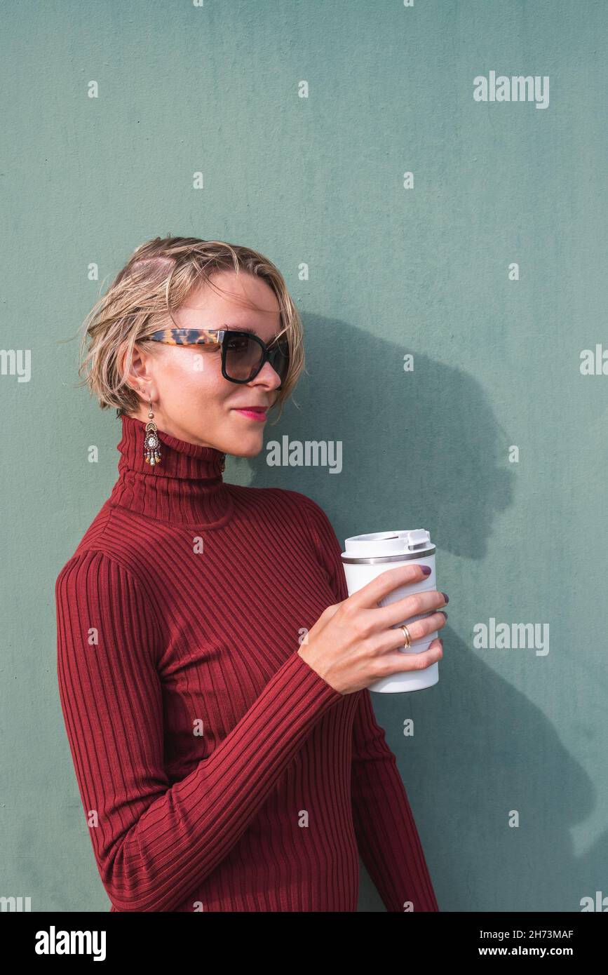 giovane donna bionda con un maglione di granata a collo di cigno che proietta l'ombra su un muro verde. Sta tenendo una tazza di caffè take away Foto Stock