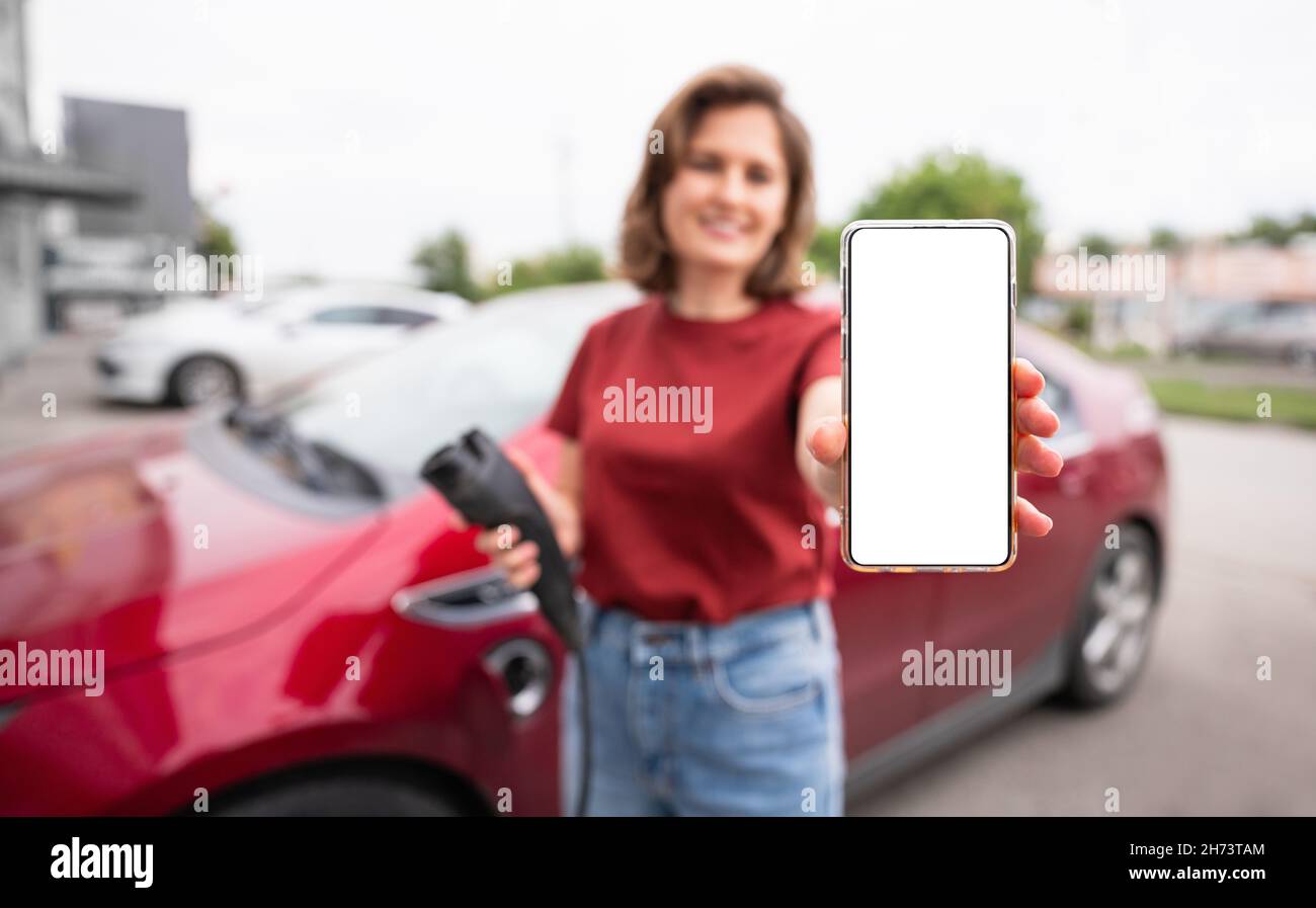 Una donna sullo sfondo di un'auto elettrica rossa mostra uno schermo vuoto dello smartphone alla fotocamera. Puoi aggiungere il tuo contenuto qui Foto Stock