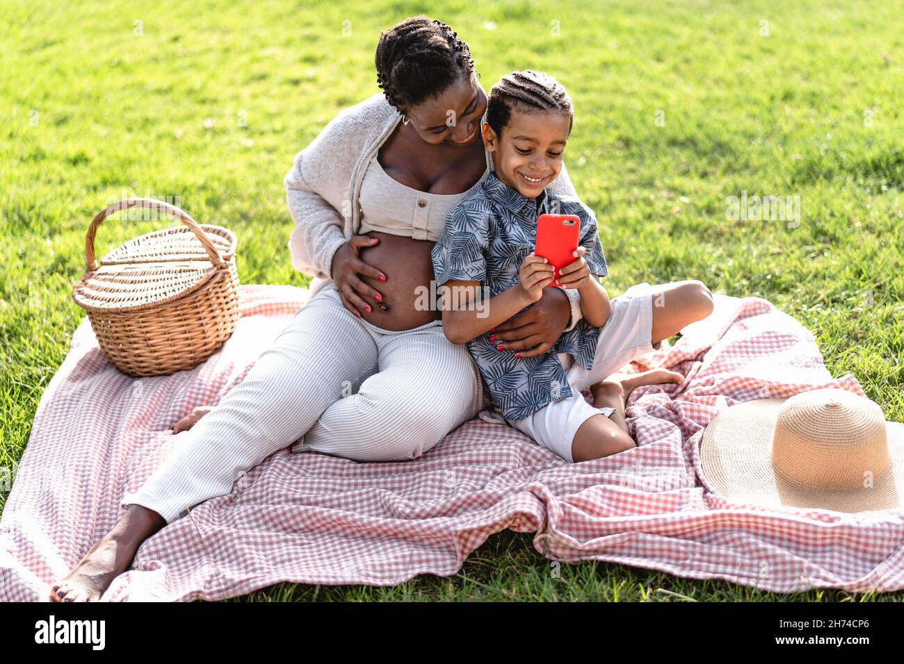 Felice madre incinta africana passare il tempo con suo figlio fare un picnic durante il fine settimana in parco pubblico - la famiglia afro guardare sullo smartphone Foto Stock