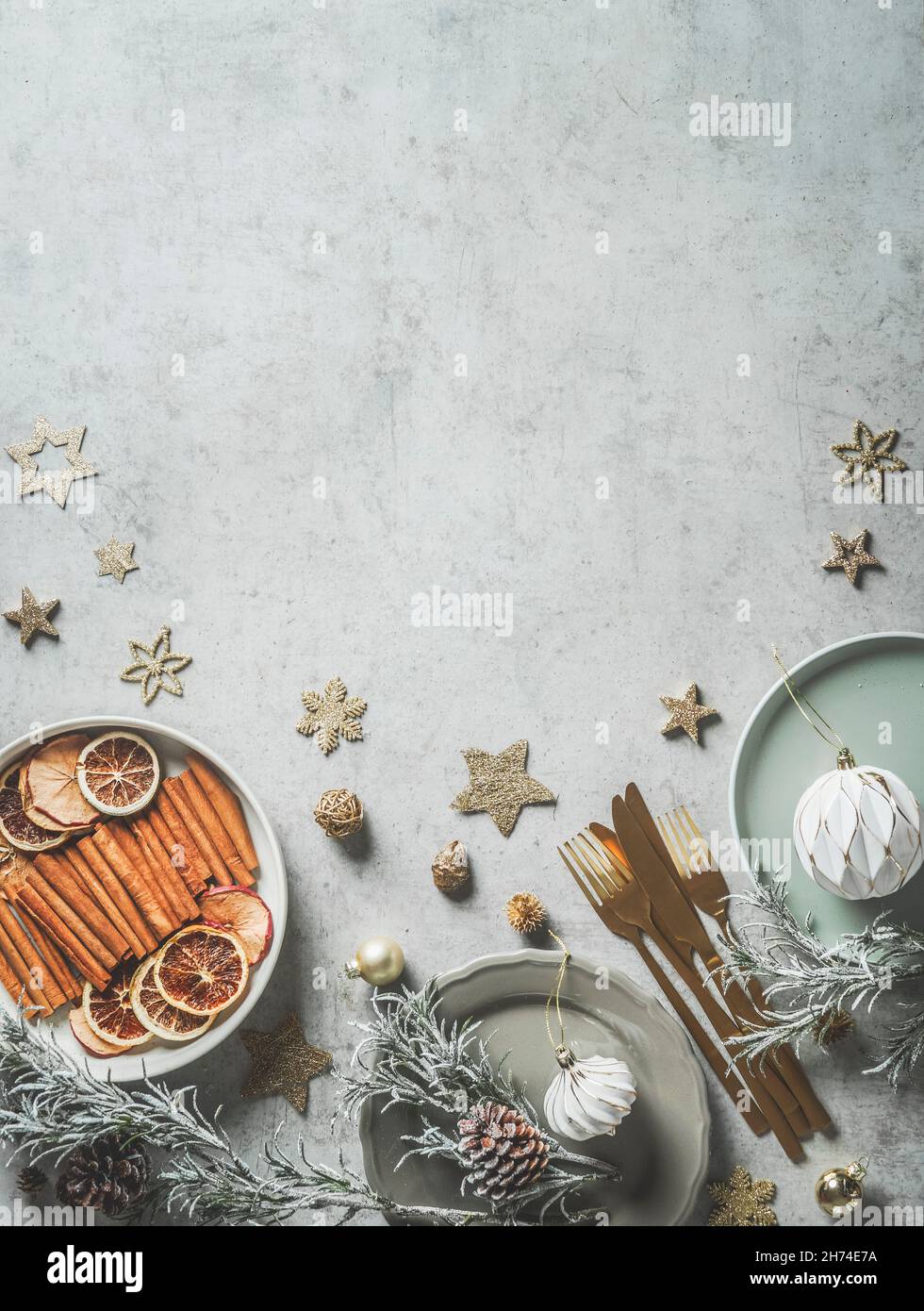 Sfondo natalizio con piatti, posate d'oro, decorazione, stelle, bastoni di cannella, fette d'arancia, bauble, coni di pino e verdi di abete su conc. grigio chiaro Foto Stock