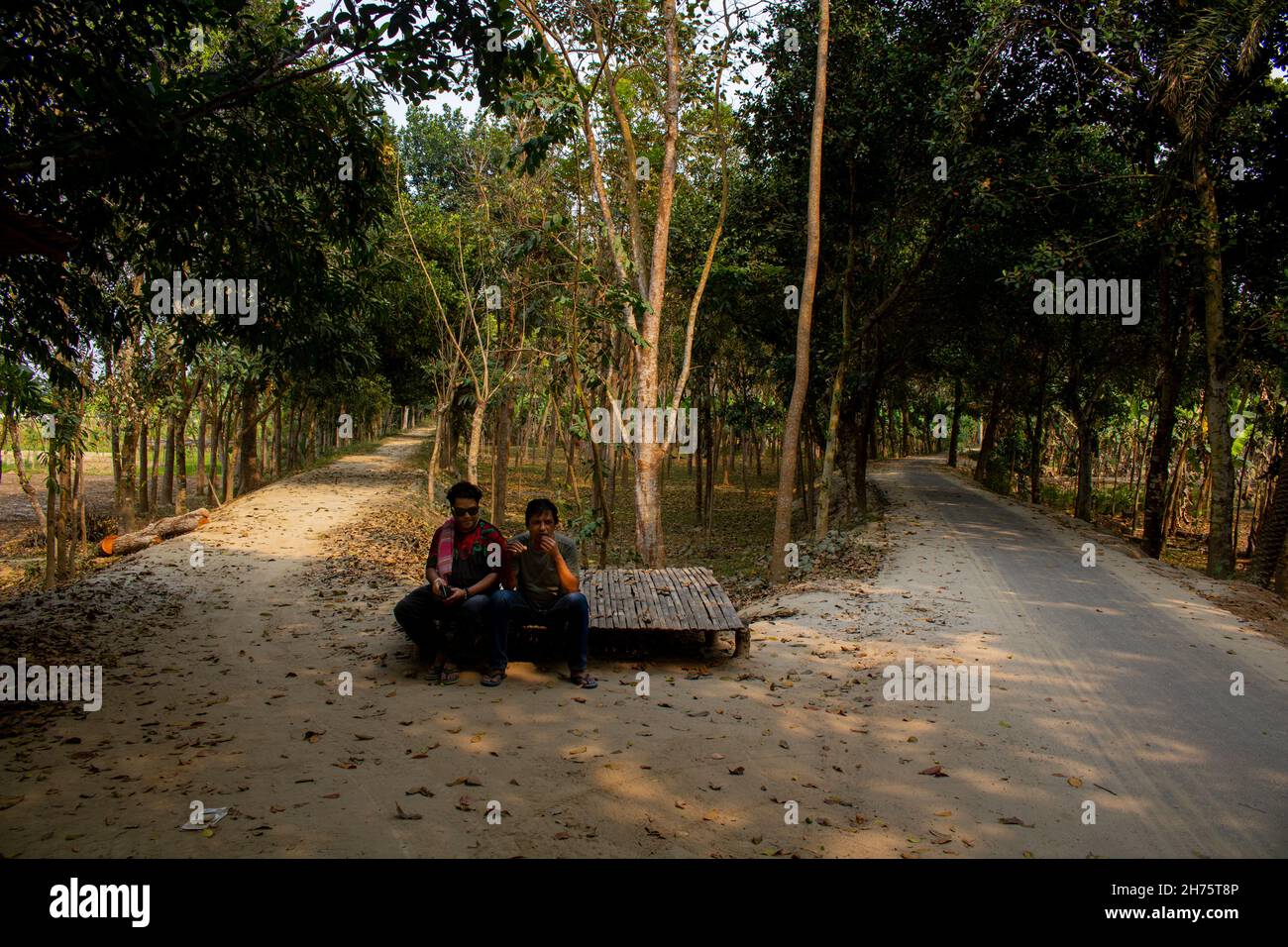 Il quadro è del contesto rurale del Bangladesh. L'ora è mezzogiorno. Due giovani uomini seduti all'ombra nel mezzo di una strada a tre vie. Foto Stock