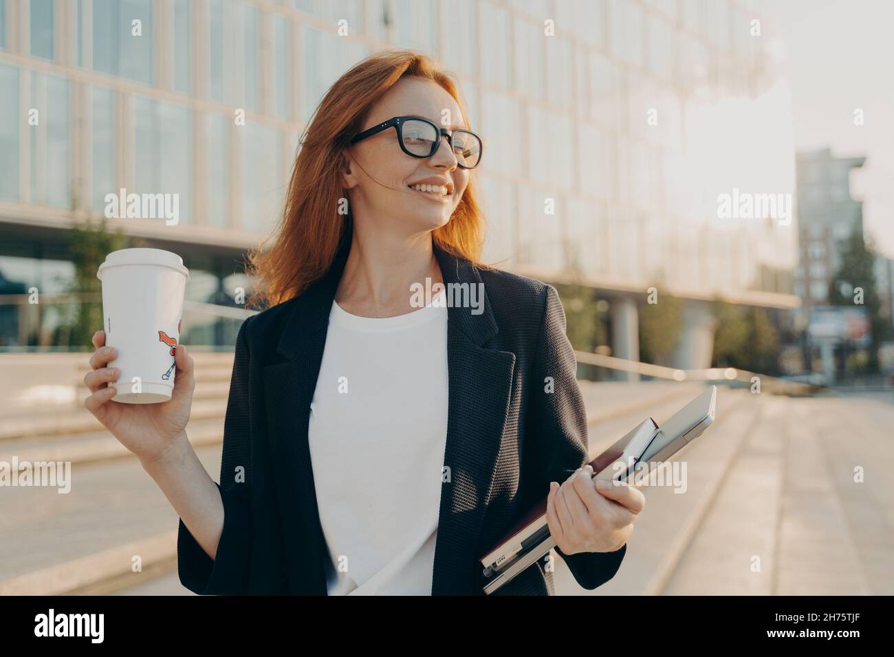 Donna allegra zenzero felice in occhiali bere caffè e tenere il computer portatile mentre si cammina all'aperto Foto Stock