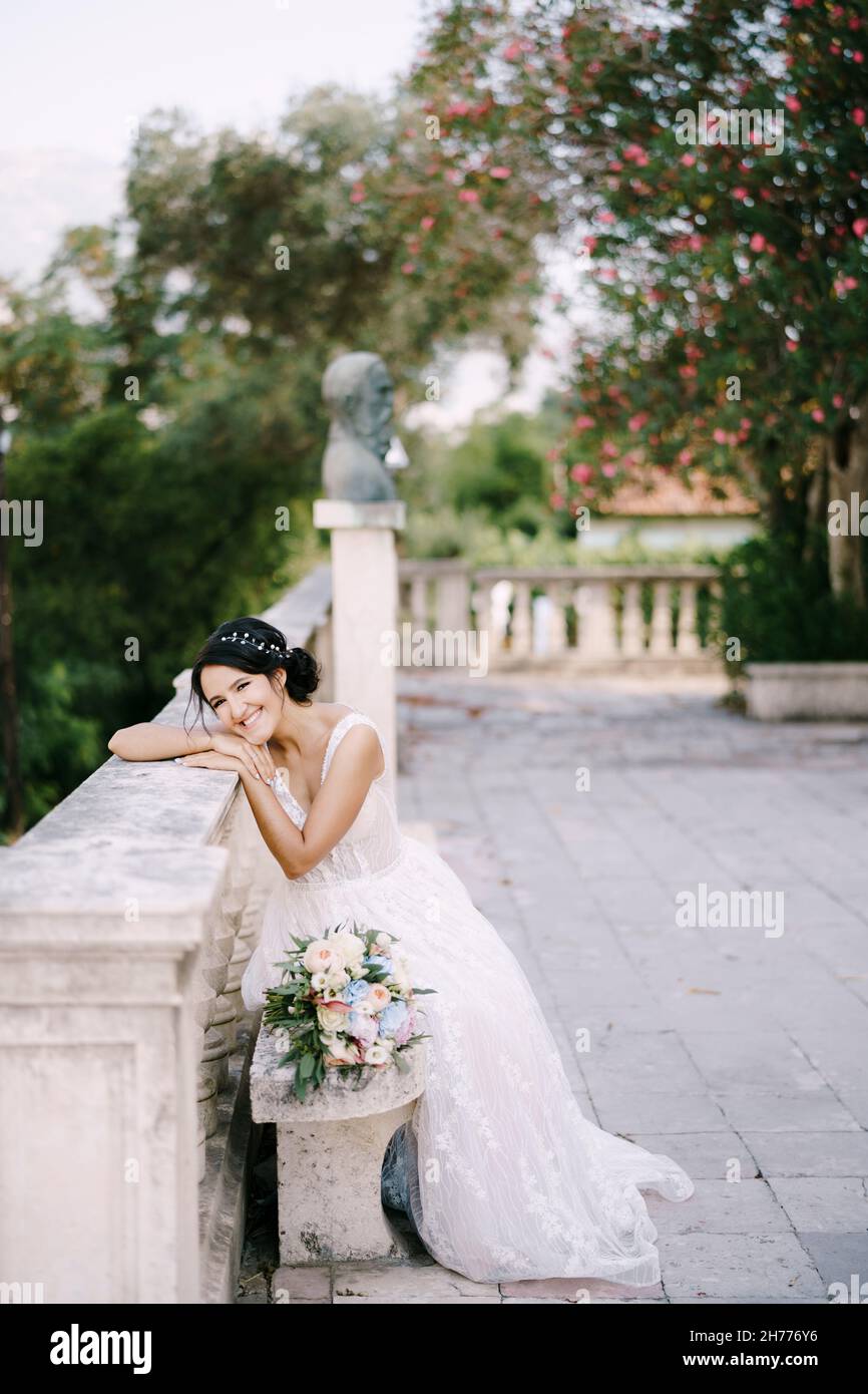La sposa si siede su una panchina su un sito panoramico nella città vecchia, si appoggia sulla ringhiera e sorride Foto Stock