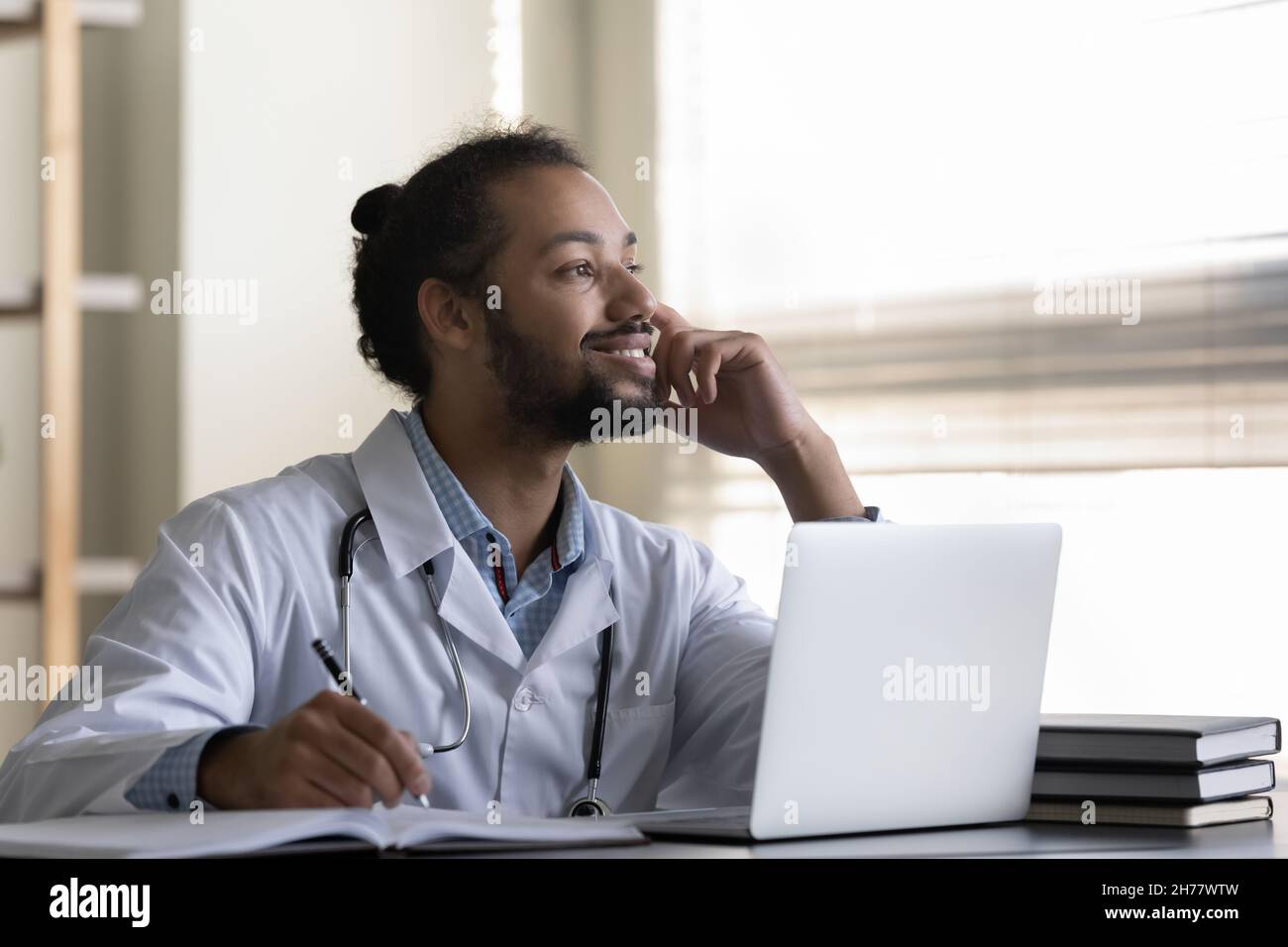Un uomo medico afro-americano che sorride pensivo prendendo appunti, usando un computer portatile Foto Stock