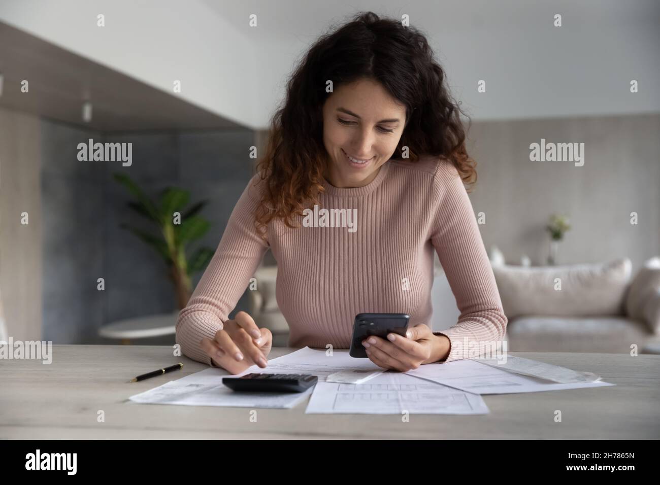 Donna sorridente con smartphone e calcolatrice, lavorando con documenti finanziari Foto Stock
