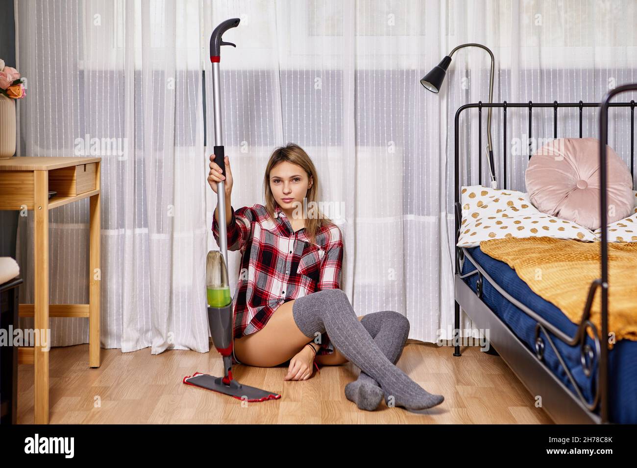 Moderno mop piatto con flacone spray e tampone in microfibra in mani di snella donna bianca di 24 anni seduta sul pavimento in camera da letto. Foto Stock