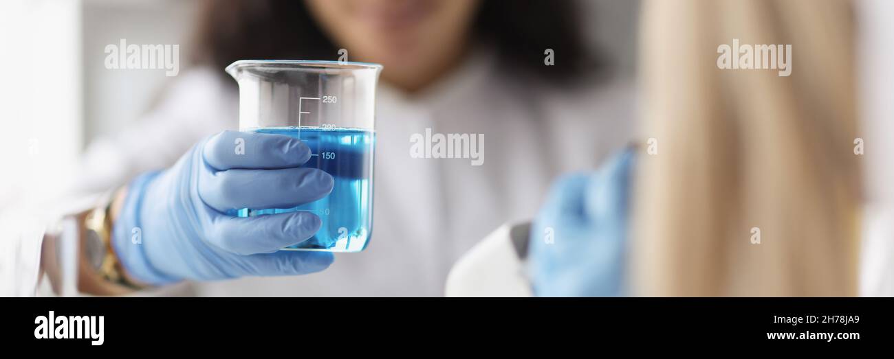 Donna chimico che tiene becher di vetro con liquido blu davanti al microscopio in laboratorio Foto Stock