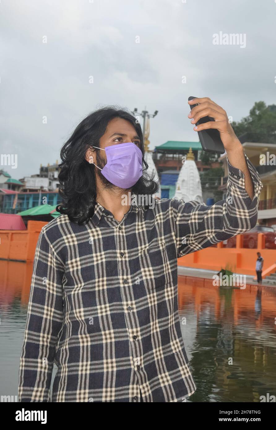 Un giovane del sud asiatico con capelli lunghi che prende selfie o che fa videochiamata con il suo smartphone con indossare maschera facciale mentre in piedi fuori Foto Stock