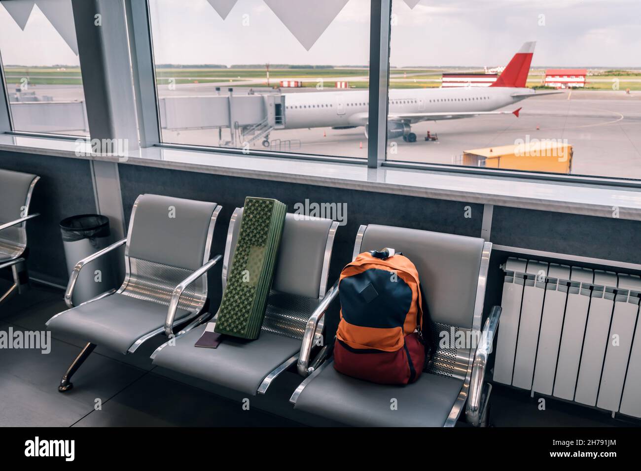 Un piccolo zaino è bagaglio a mano su una panca con vista sul cancello e un aereo parcheggiato. I passeggeri sono a bordo. Foto Stock