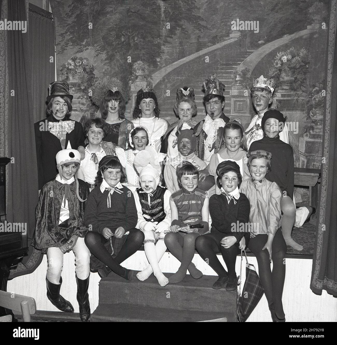 1965, storico, seduto insieme su un piccolo palco, sorrisi da un gruppo di bambini di un centro giovanile vestito nei loro costumi per il gioco, Alice in Wonderland, Fife, Scozia, Regno Unito. Foto Stock