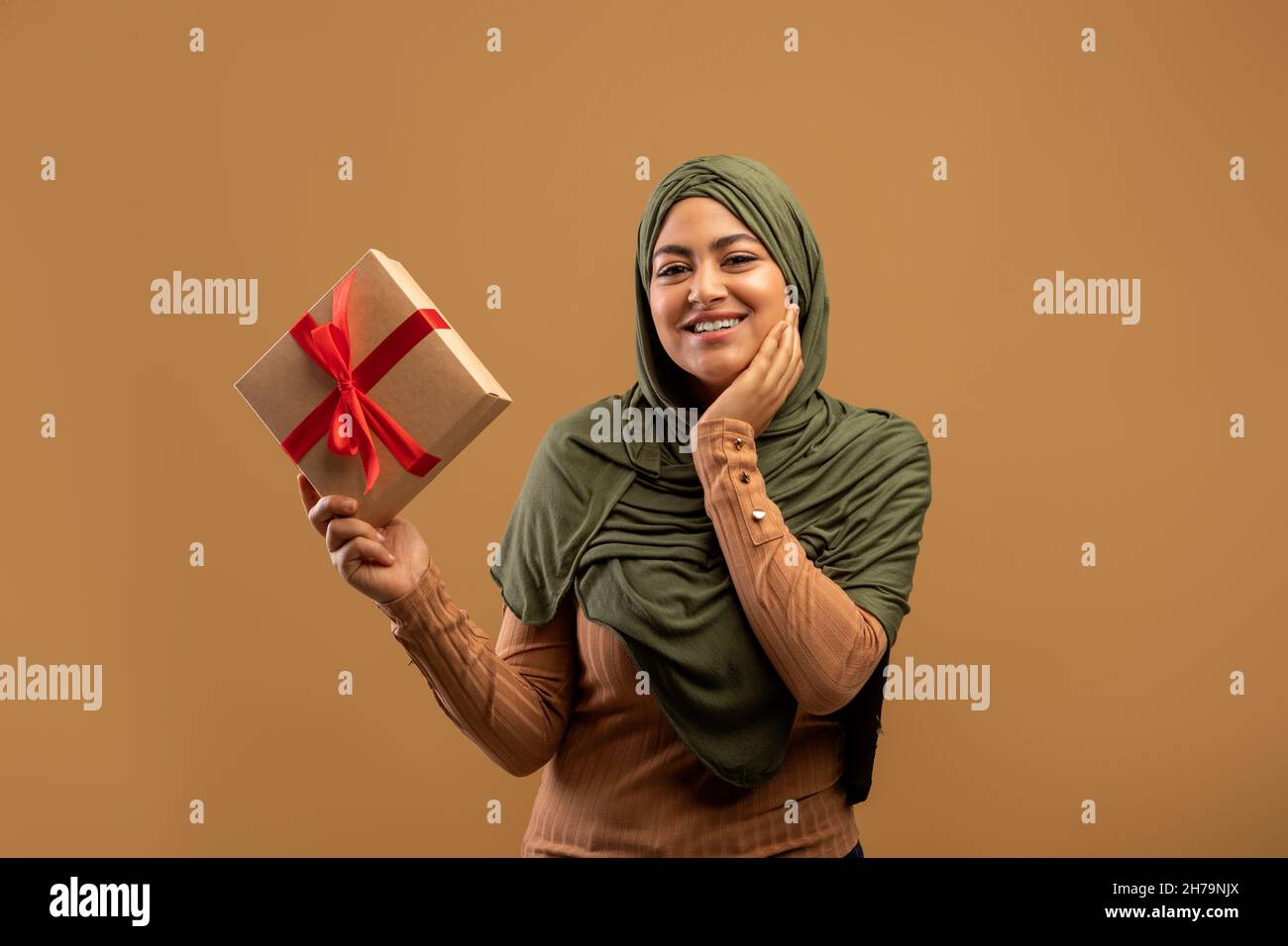 Signora musulmana felice in hijab che tiene la scatola avvolta, ritenendosi eccitato su sfondo marrone, ricevendo la sorpresa di compleanno Foto Stock