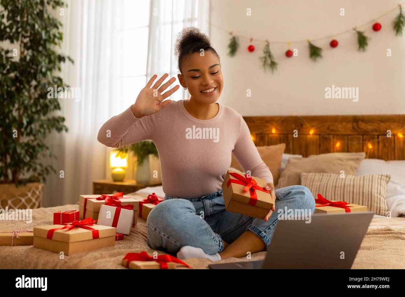 Positivo afroamericana signora che offre la scatola del regalo al webcam del laptop, videochiamata sul laptop, celebrando il Natale Foto Stock