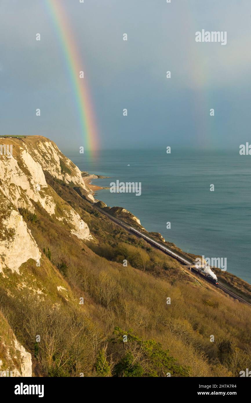 La locomotiva a vapore Clan Line corre lungo la costa a Cautle-le-ferne tra dover e Folkstone con il Belmond British Pullman sotto un arcobaleno autunnale Foto Stock