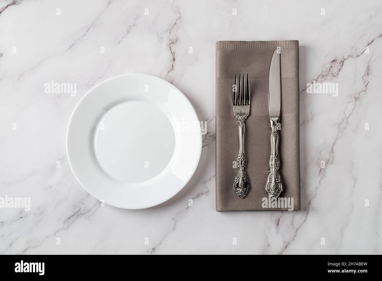 Piatto in porcellana bianca vicino all'elegante coltello da tavolo vintage e forchetta su un tovagliolo grigio da pranzo sopra il tavolo in marmo. Impostare la tabella. Svuotare la piastra in ceramica pulita. Foto Stock