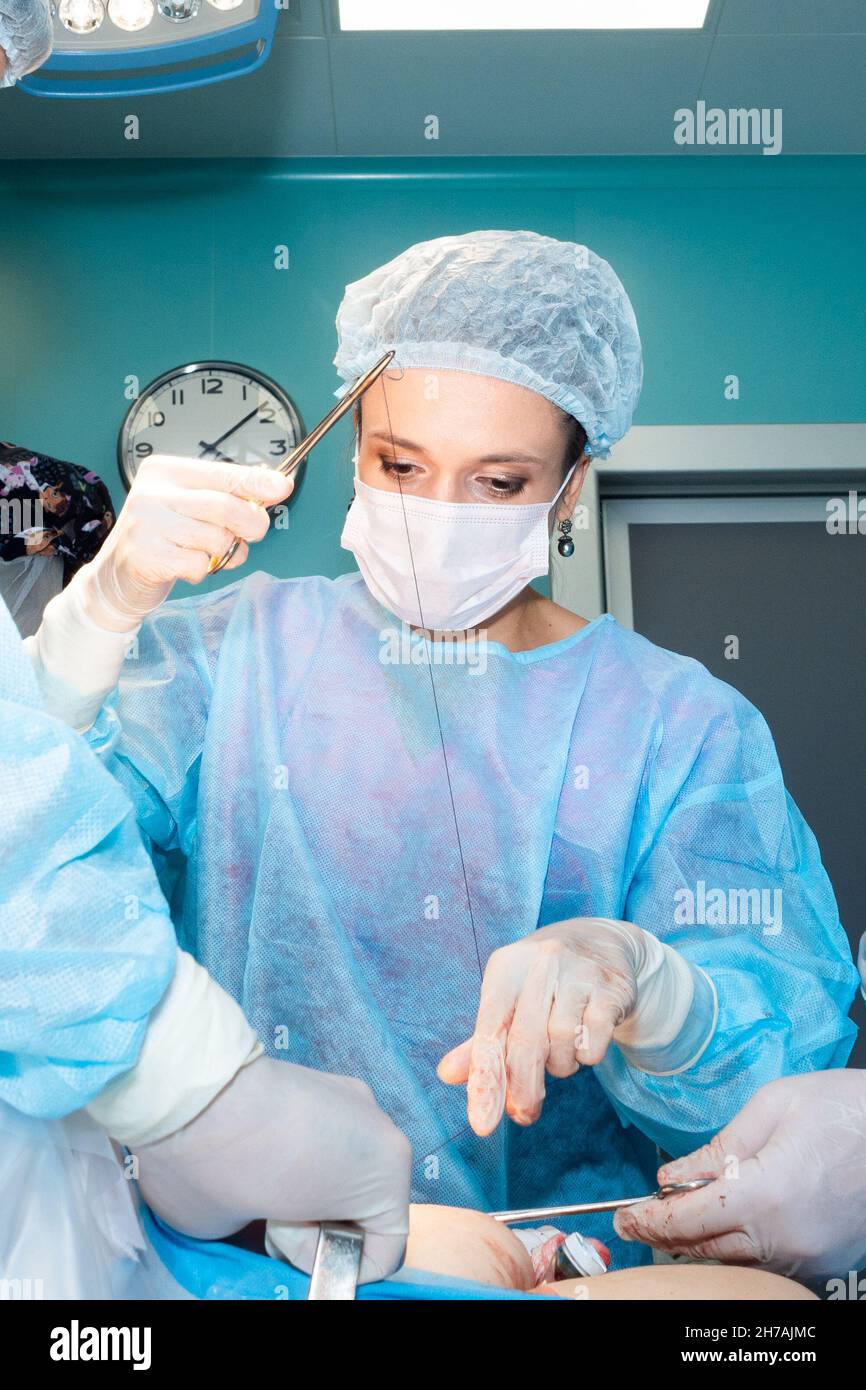 Un chirurgo femminile sutura la pelle del paziente con un supporto dell'ago e sutura chirurgica. Messa a fuoco selettiva. Chirurgo che indossa guanti sterili, uniforme chirurgica e maschera chirurgica. Foto Stock