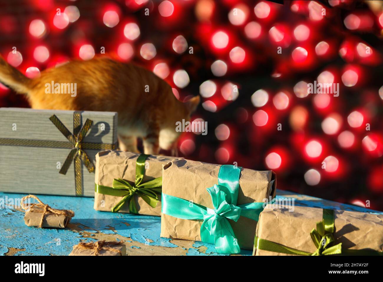 Gatto zenzero scappare vicino mestiere avvolto scatole regalo vintage su sfondo blu vecchio legno. Presenta la scatola in carta artigianale con arco verde con ombra Foto Stock