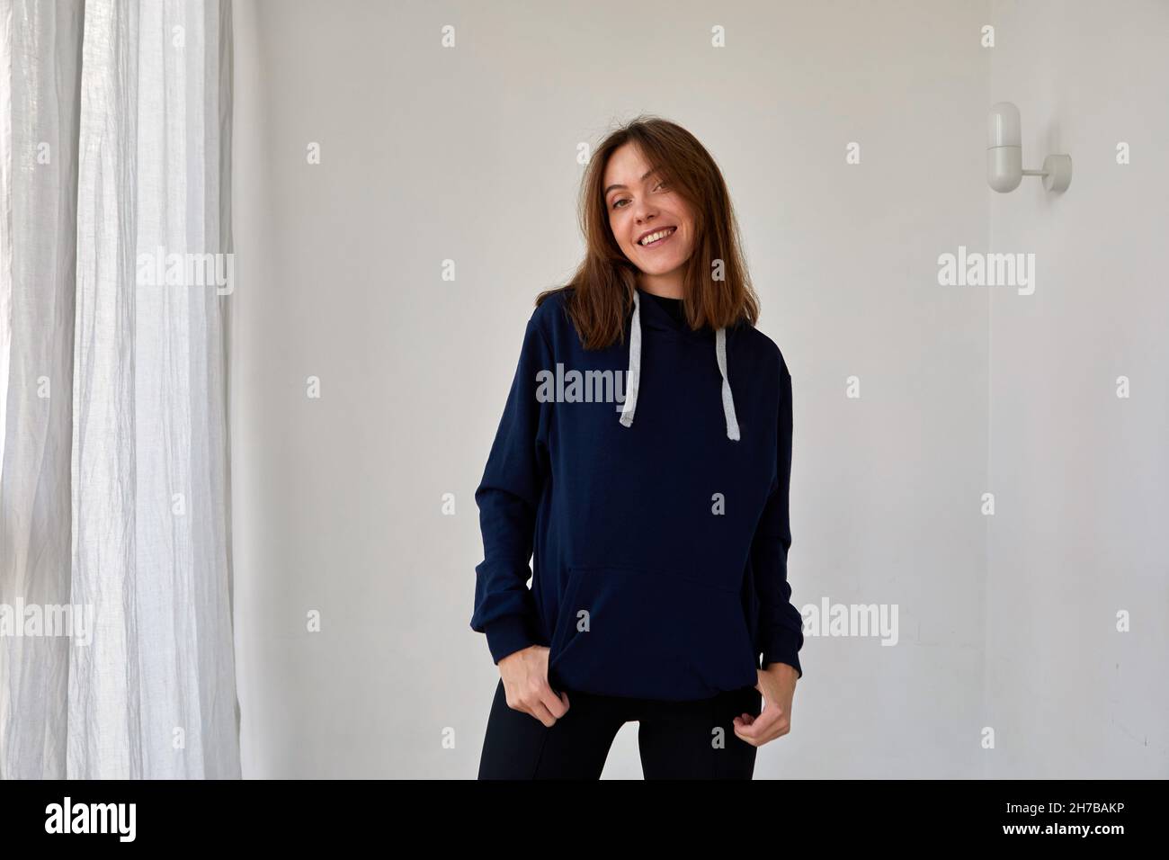 Positiva femmina in elegante felpa con cappuccio in piedi in stanza con pareti e tende bianche e guardando la macchina fotografica Foto Stock