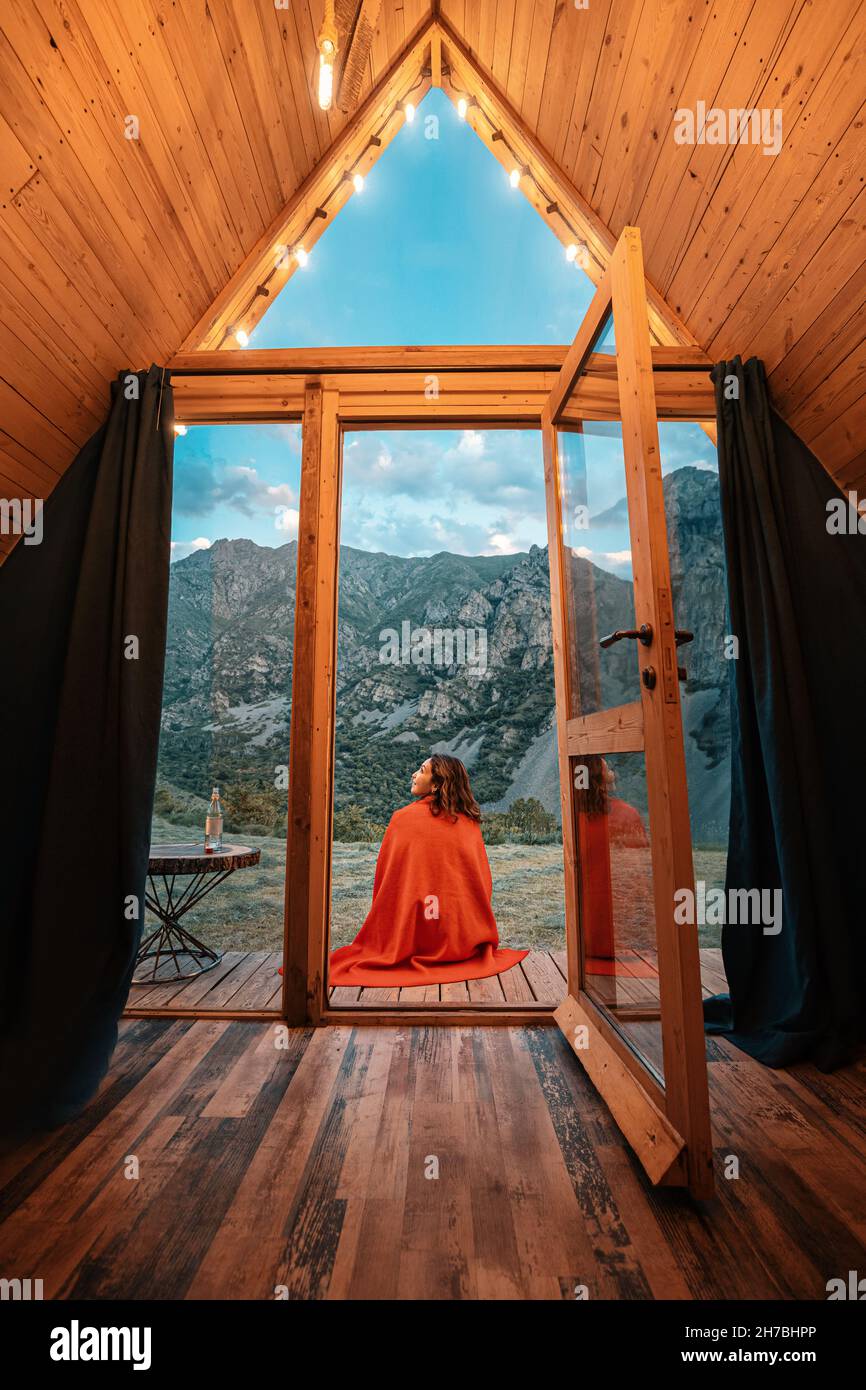 Donna sola sul portico di una piccola casa di legno in montagna. Il concetto di glamping e vacanze idilliache Foto Stock