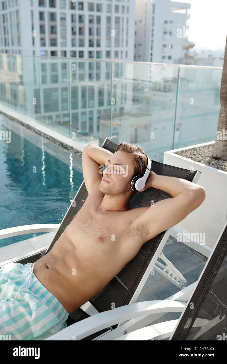 Bel giovane uomo che ascolta la musica in cuffia e si appoggia indietro in chaise-lounge presso la piscina sul tetto Foto Stock
