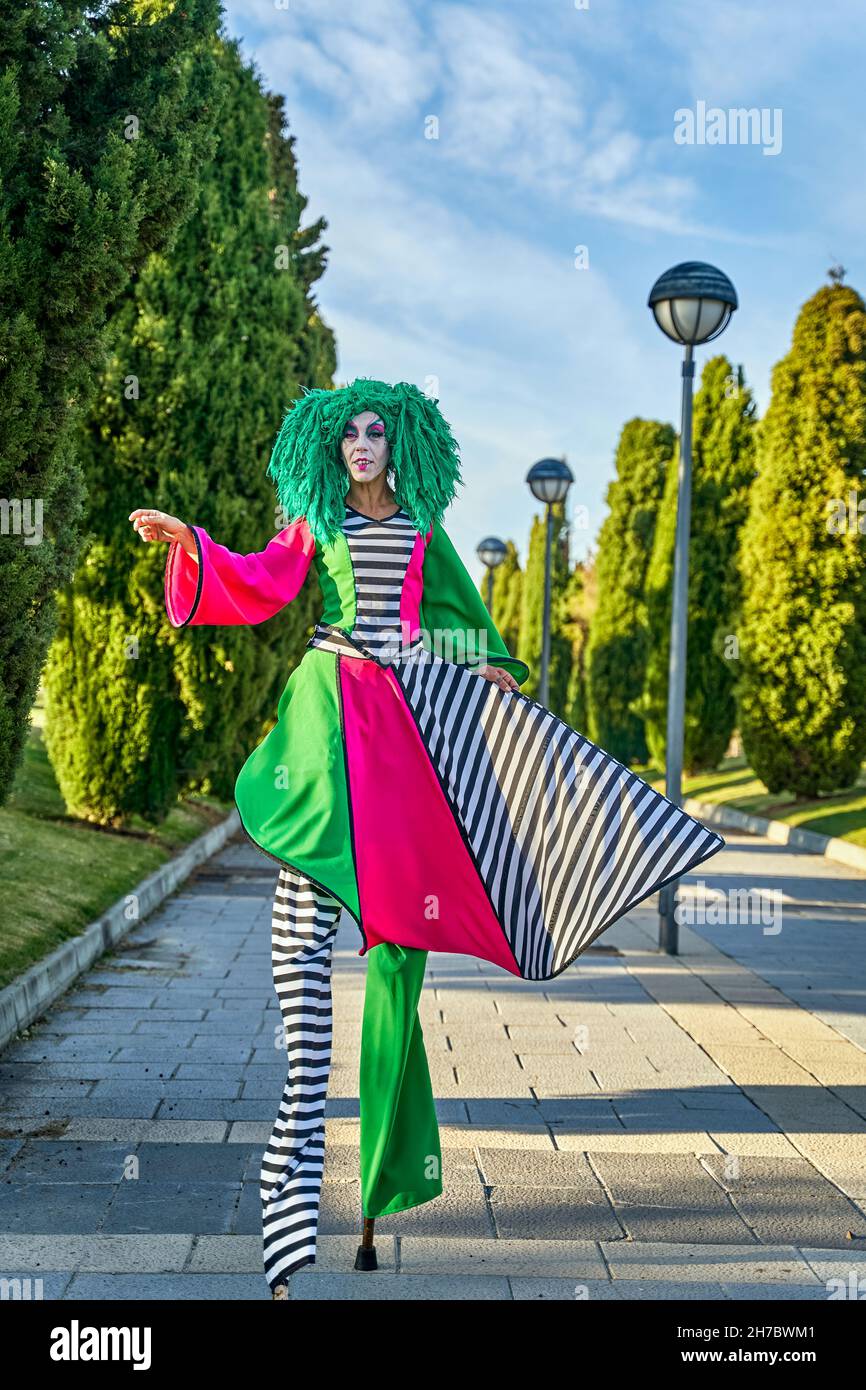 Un'intera serie di camminatori a palafitte femminili in un divertente costume da harlequin e una parrucca verde che passeggia sul vicolo tra alberi lussureggianti durante la sfilata nel parco soleggiato Foto Stock