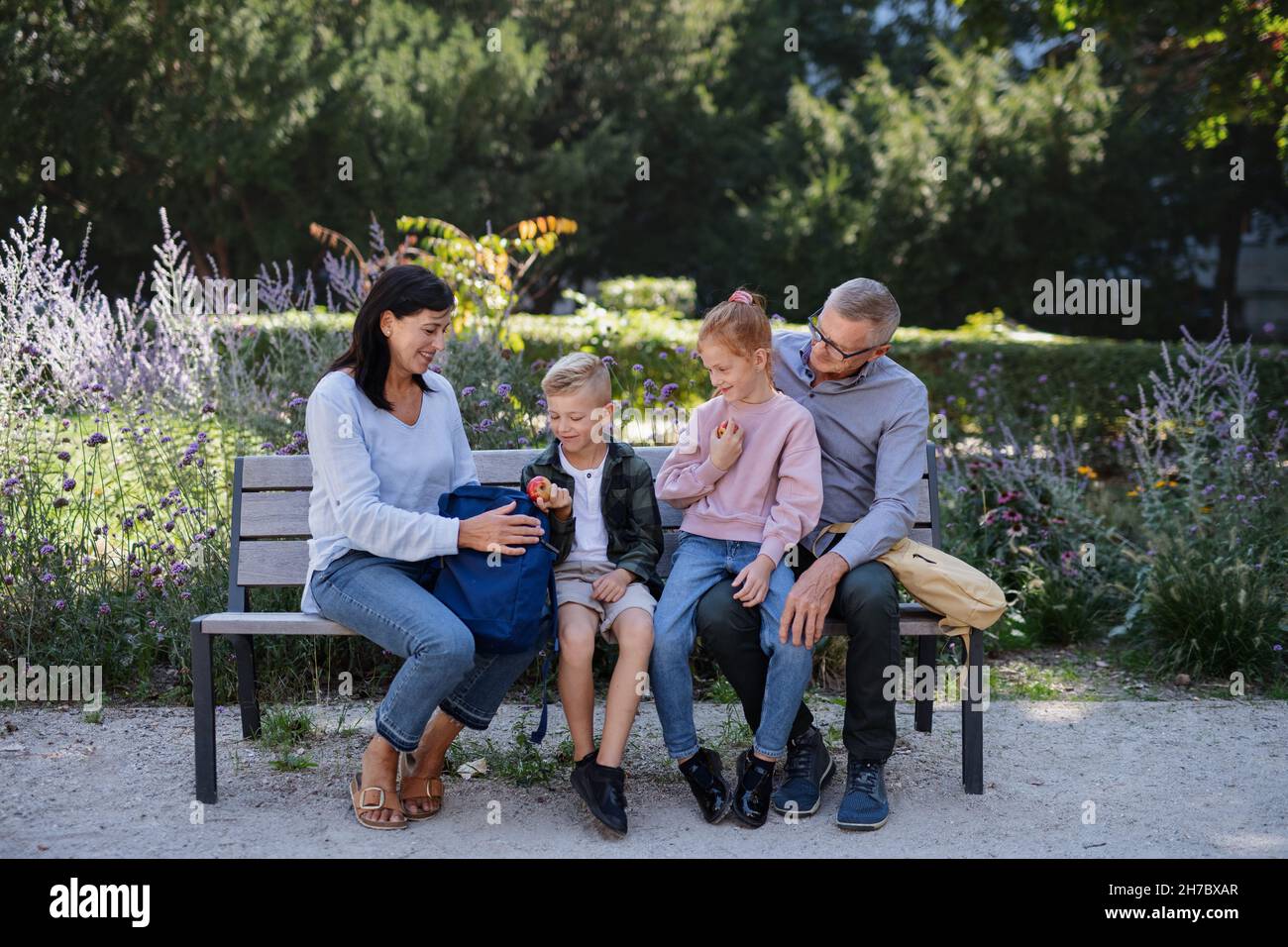 Coppia senior con nipoti seduti su panca, mangiando snack e parlando all'aperto nel parco. Foto Stock