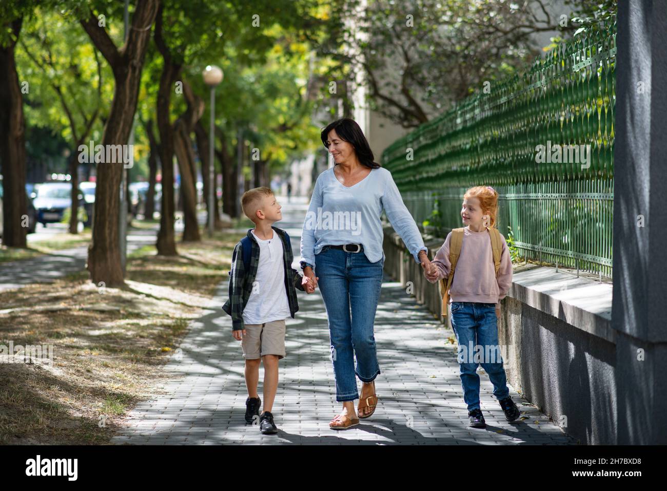 Nonna felice che porta i nipoti a casa dalla scuola, camminando all'aperto in strada. Foto Stock