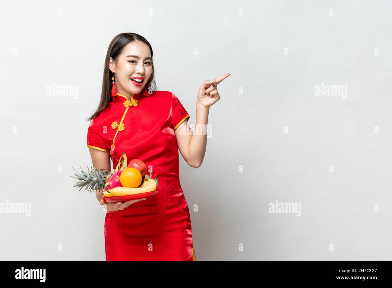 Ritratto cinese di Capodanno di donna asiatica sorridente in abito tradizionale gheongsam rosso con frutti fortunati che punta la mano allo spazio vuoto in isolat grigio chiaro Foto Stock