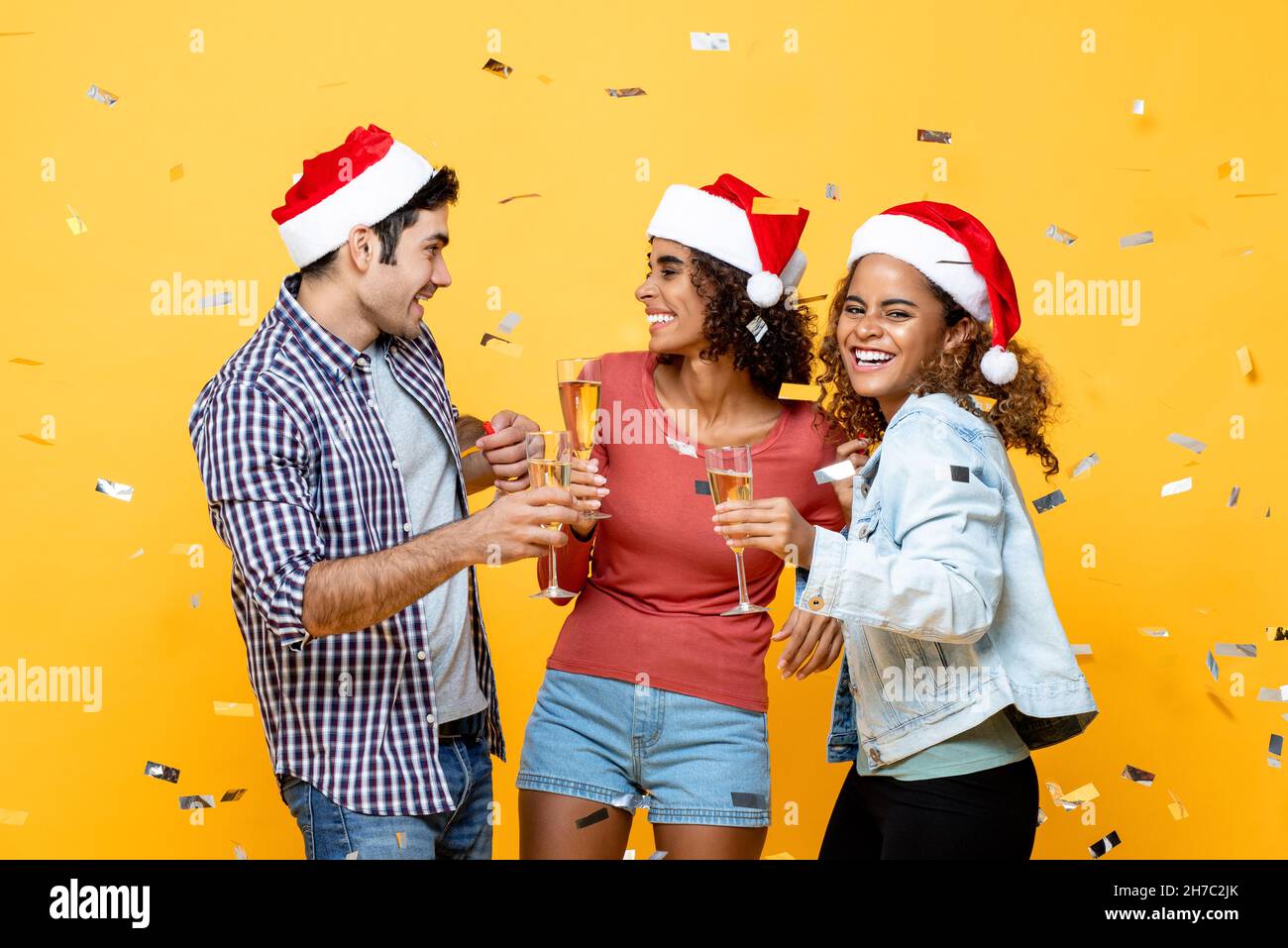Ritratto di tre amici allegri e diversi che bevono champagne mentre festeggiano il Natale su sfondo giallo dello studio con confetti Foto Stock
