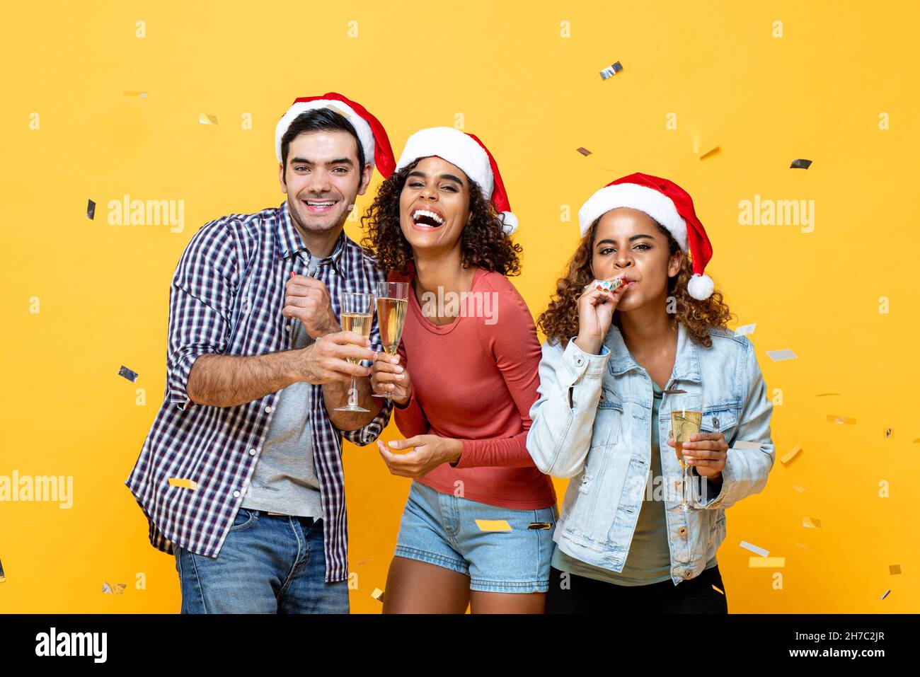 Ritratto del gruppo di amici allegri e variegati con champagne che festeggia il Natale su sfondo giallo dello studio con confetti Foto Stock