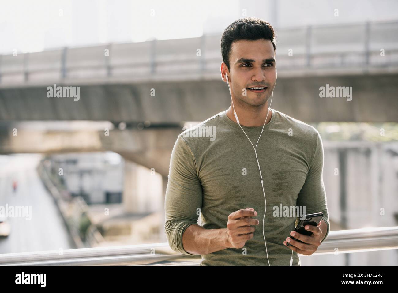 Sorridente giovane sudato bell'uomo indiano che ascolta musica su smartphone all'aperto in città dopo il jogging Foto Stock