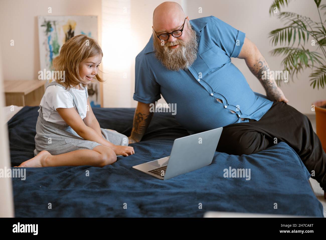Il padre bearded e la bambina osservano la lezione di moovie via il laptop moderno che si siede sul letto Foto Stock