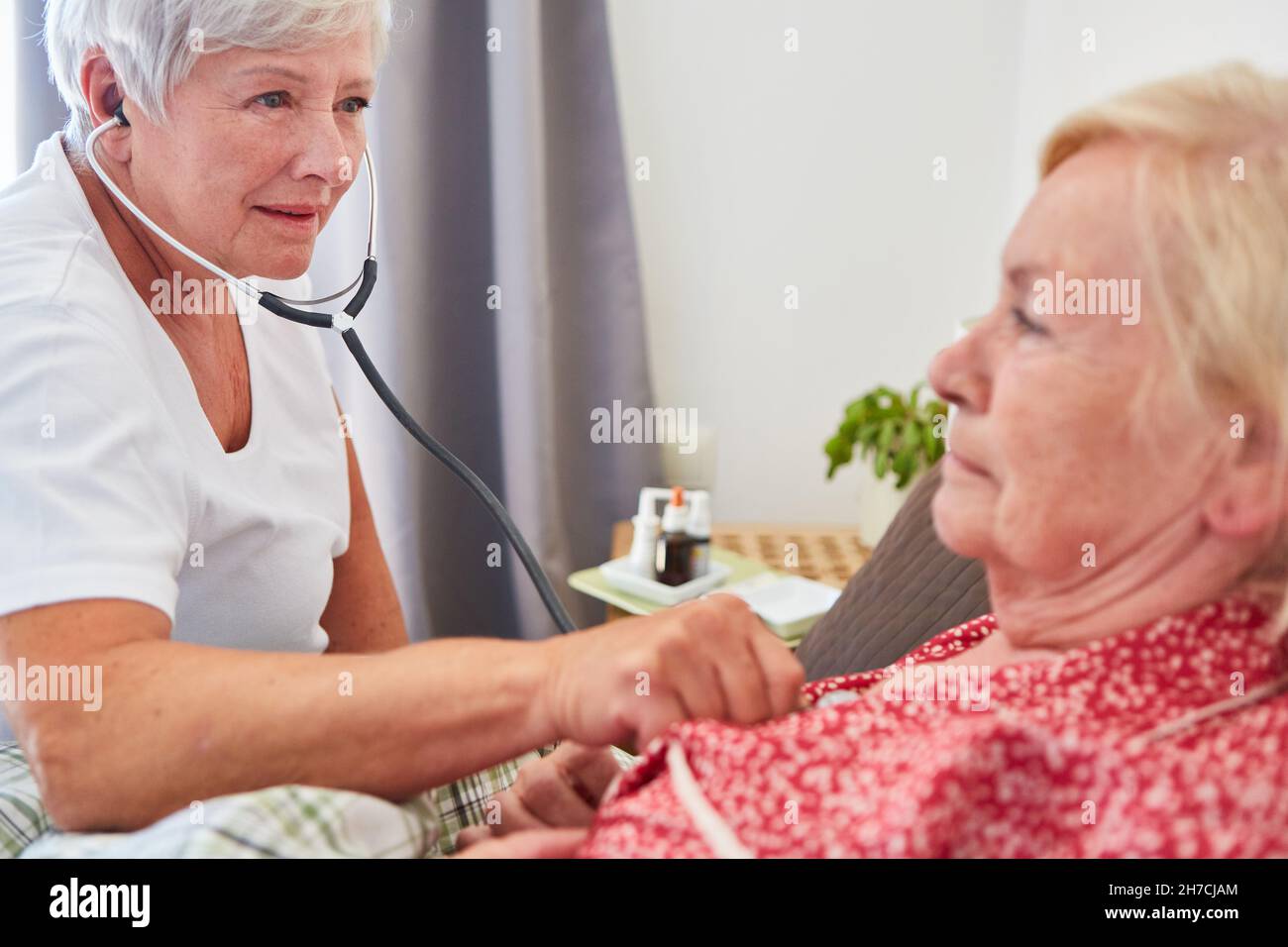 Un medico o un infermiere geriatrico esamina una donna anziana a letto in una casa di riposo con uno stetoscopio Foto Stock